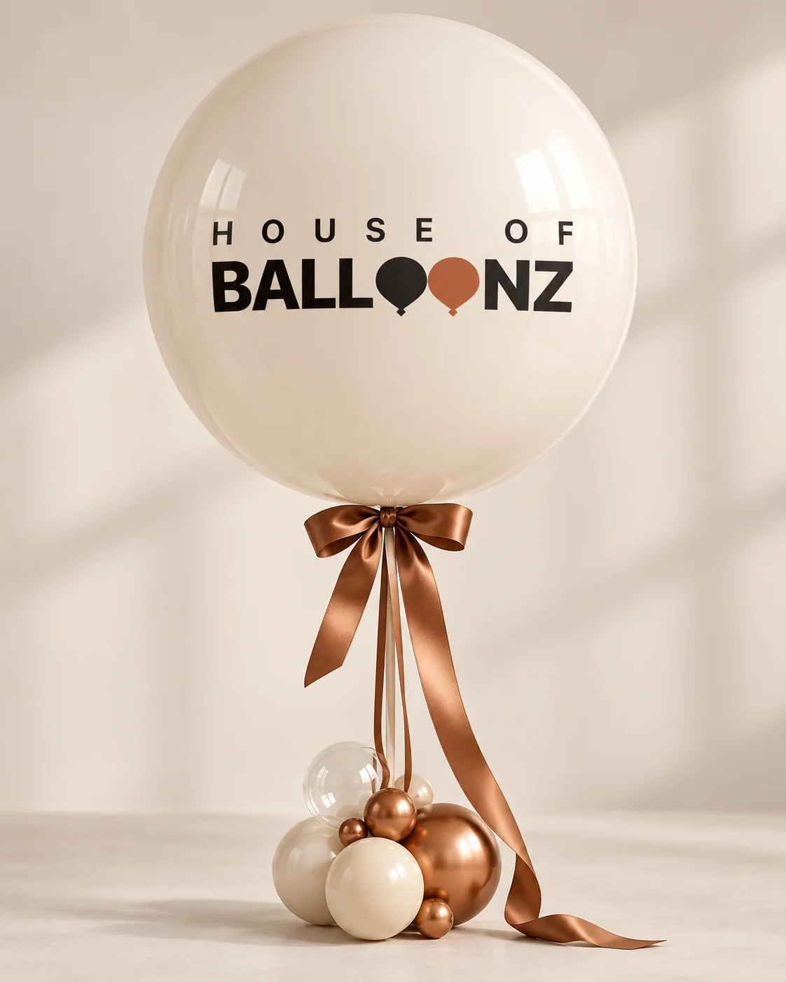 A large cream House of Balloonz balloon tied with a copper ribbon, above a cluster of cream and copper balloons.