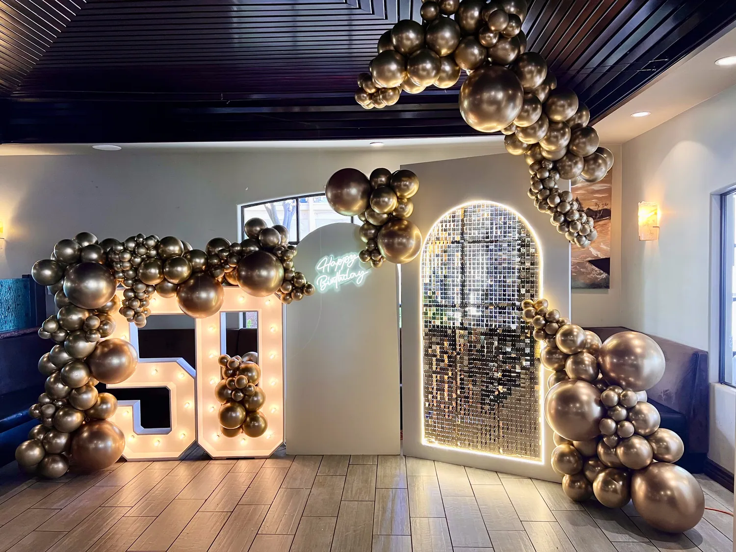 Gold chrome balloon garland running across a restaurant ceiling to a light-up 50 and a gold sequin wall.