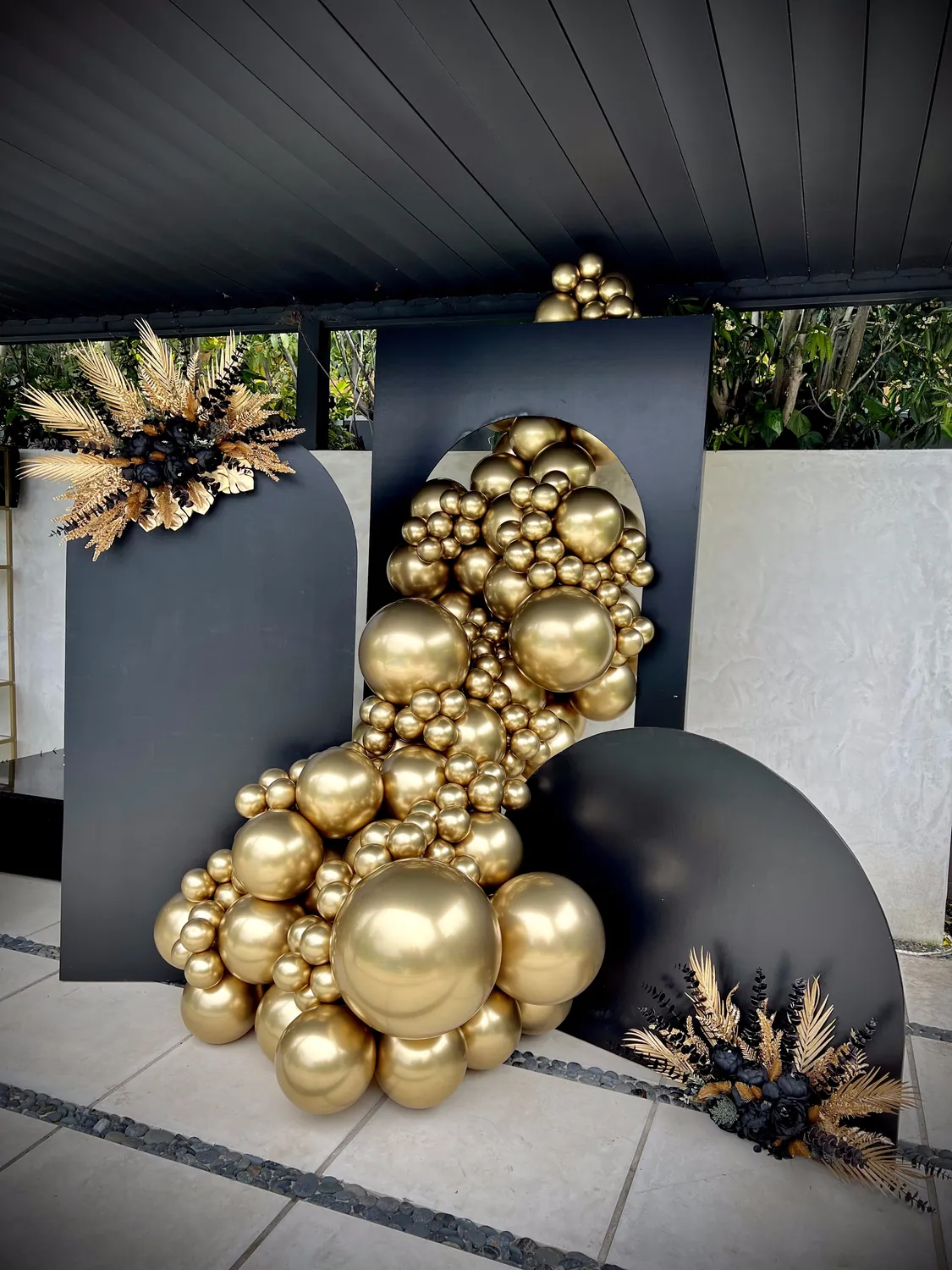 Gold chrome balloon cluster on charcoal panels with dried palm fans, on a patio.