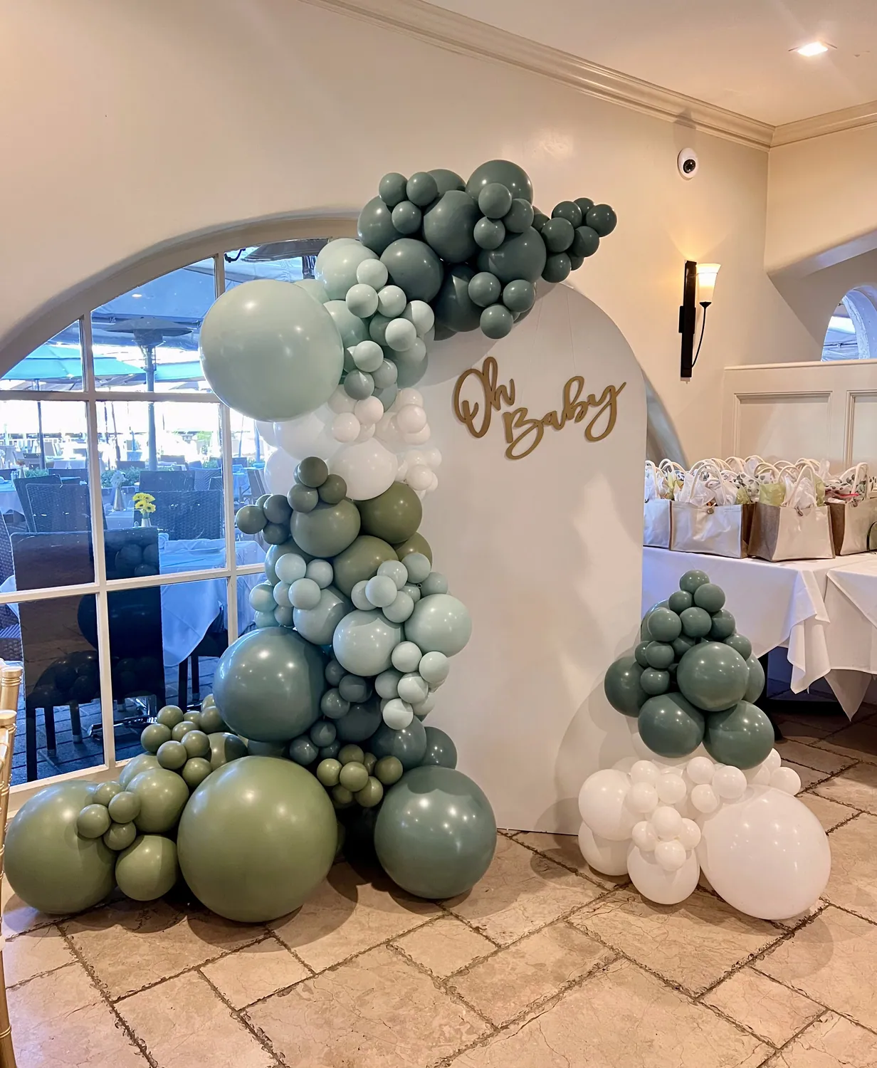 Sage green and white balloon backdrop with an Oh Baby sign in a waterfront dining room.