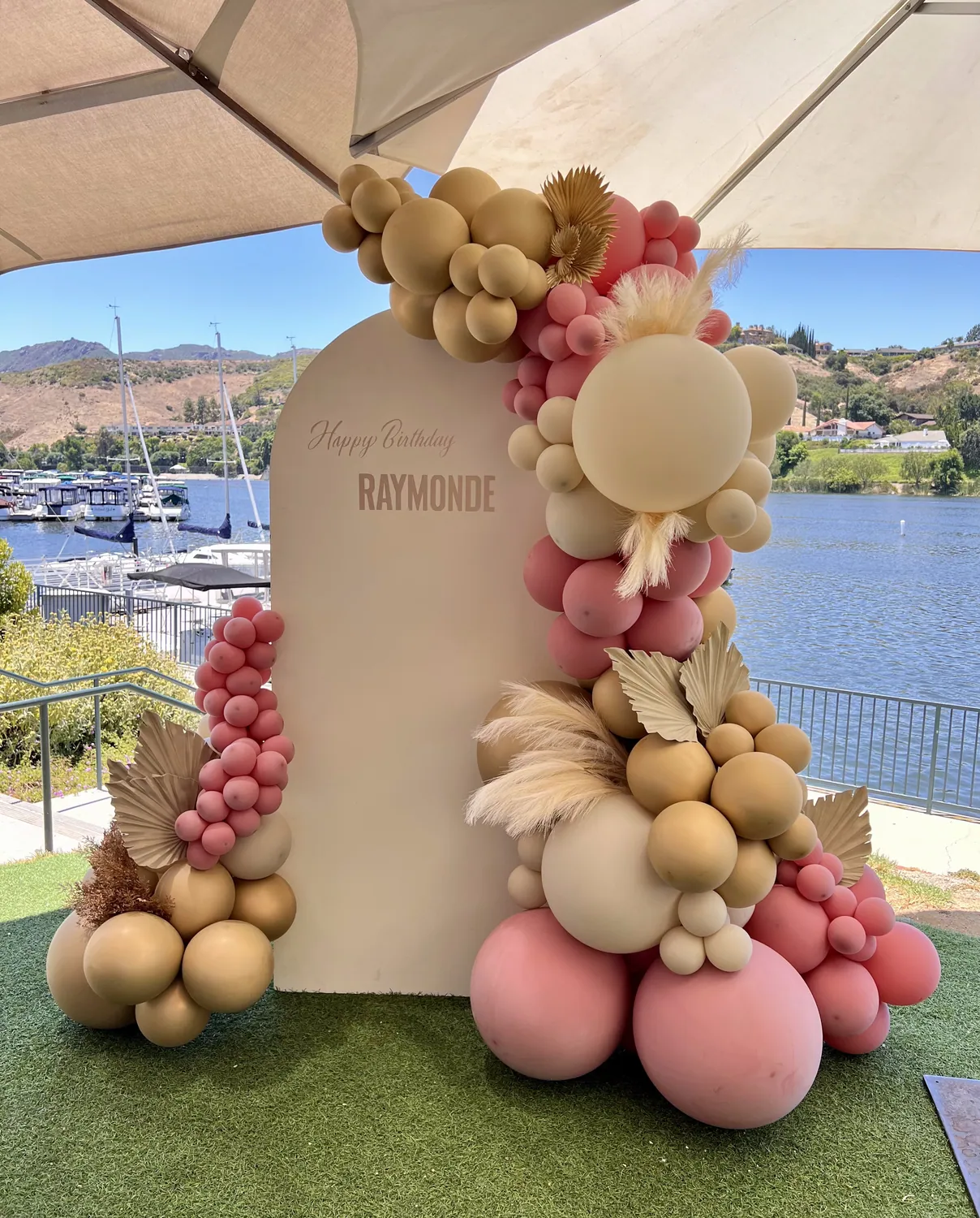 Rose, sand and gold balloon backdrop with pampas against an arched panel beside a marina.
