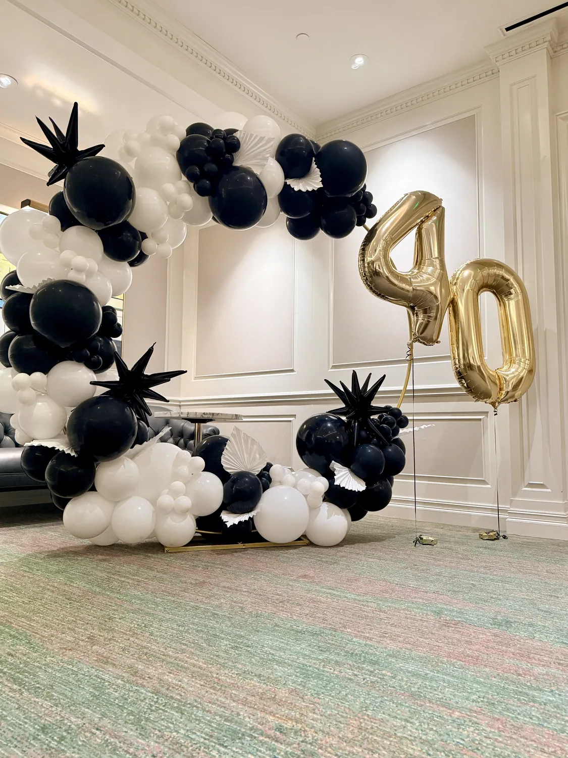 Black, white and gold balloon arch with foil starbursts and a gold 40 in a panelled room.