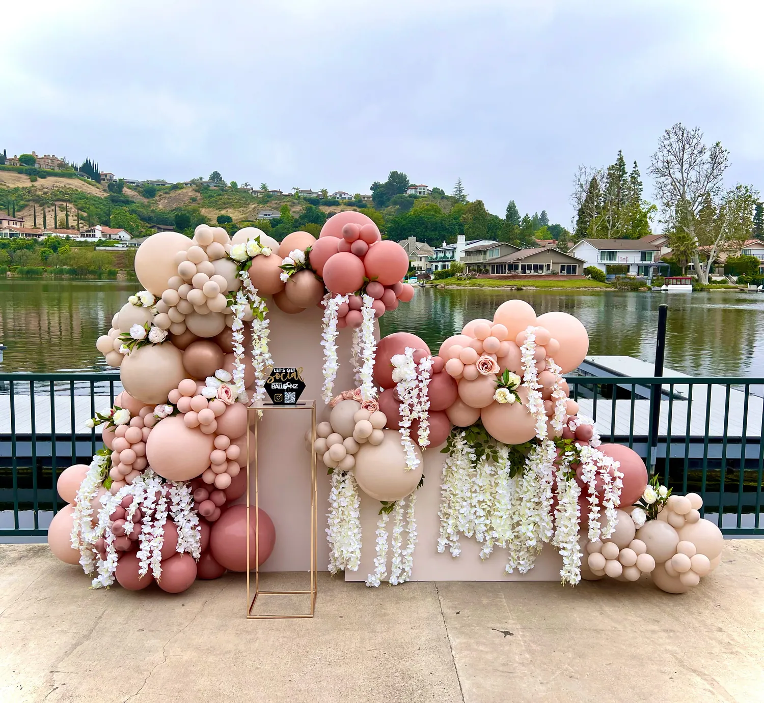 Blush and sand balloon backdrop with hanging white wisteria on a terrace above a lake.