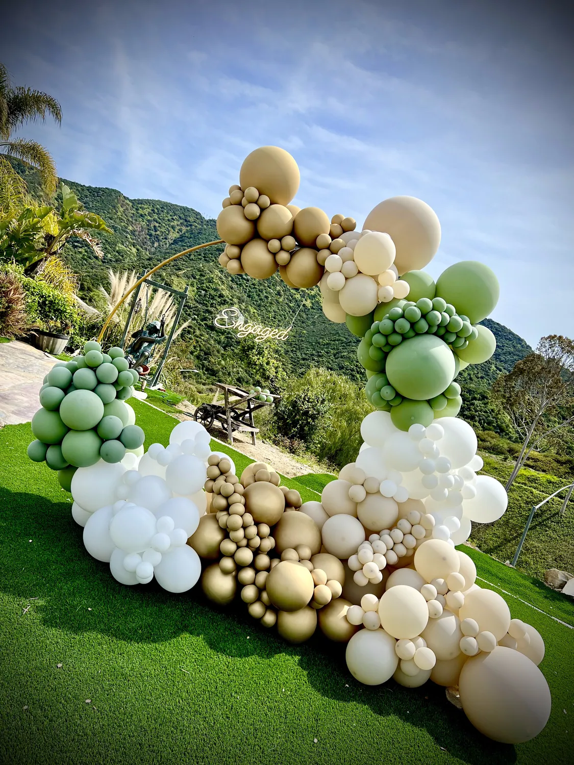Sage green, cream and gold balloon garland curving across a lawn with hills behind.