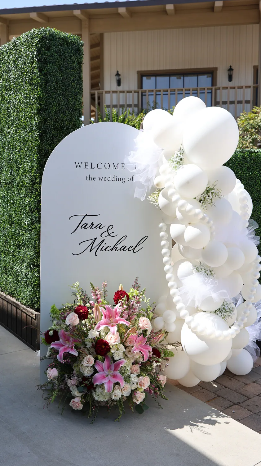 Tall white balloon arch with florals framing a wedding welcome sign beside a hedge wall.