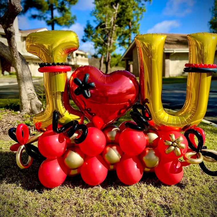 Giant gold foil letters spelling I love you with a red balloon cluster on a lawn.
