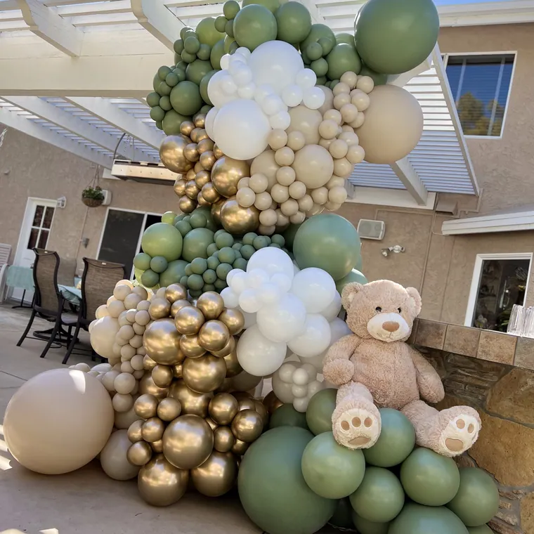 Sage, cream and gold balloon garland with a teddy bear on a patio.