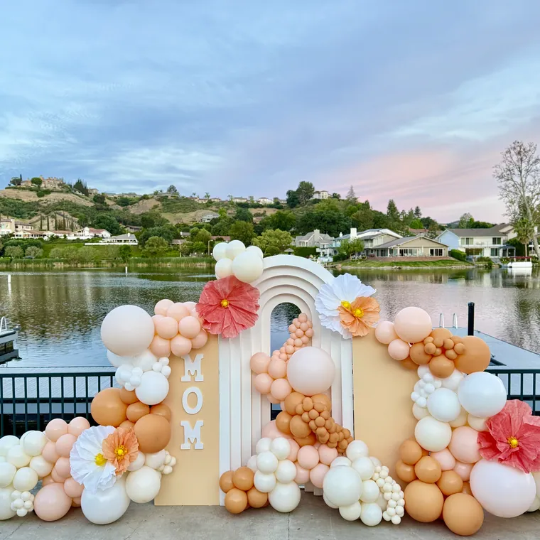 Cream and peach balloon backdrop with a rainbow arch and MOM lettering above a lake.