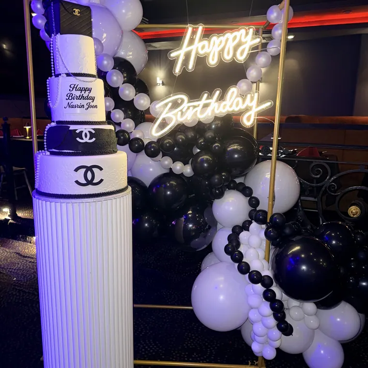 Tall white and black balloon column with a neon Happy Birthday sign behind it.