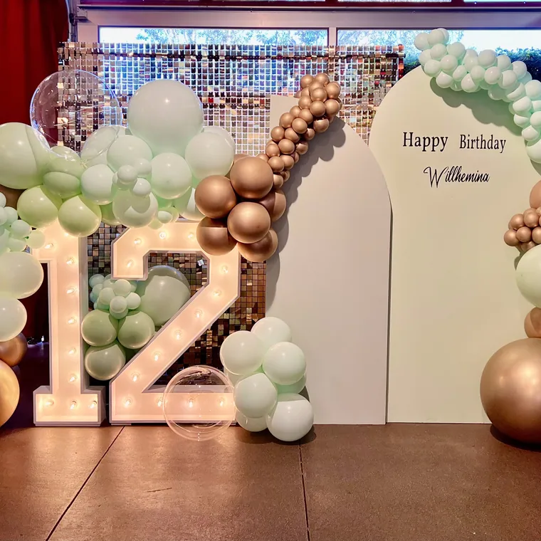 Light-up marquee number 12 beside a sage and copper balloon garland.