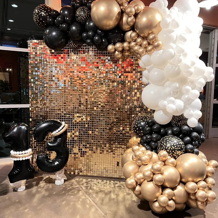 Gold sequin wall framed by a black, white and gold balloon garland.
