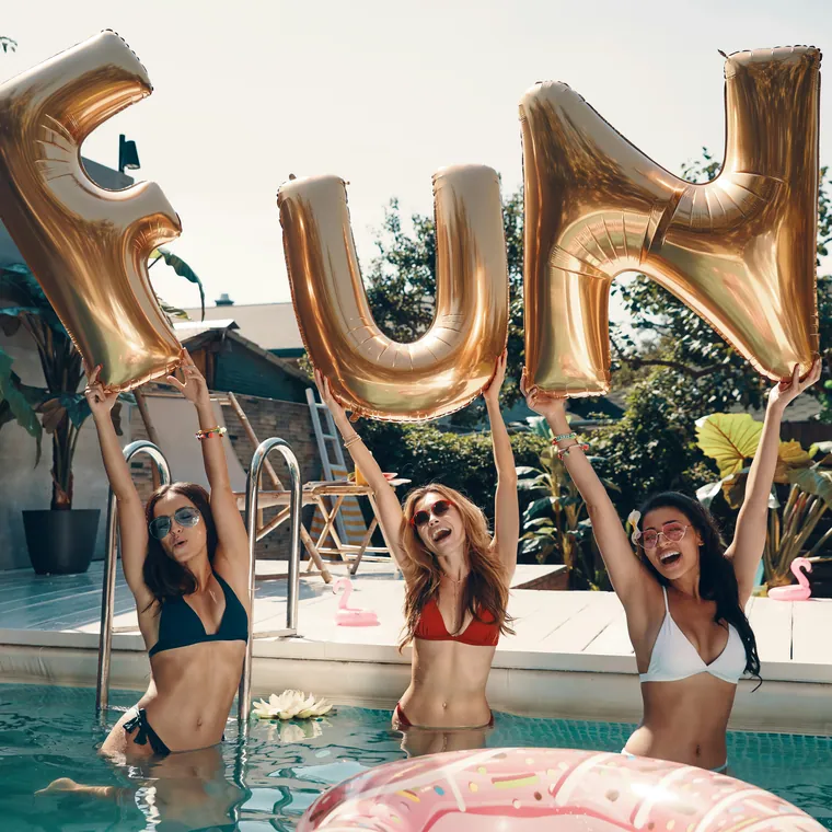 Oversized gold foil letters spelling FUN held up beside a swimming pool.