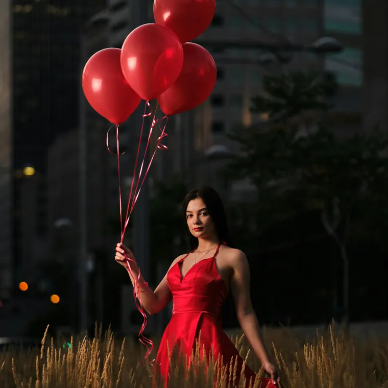 Woman in a red dress holding a cluster of red balloons with a city skyline behind.