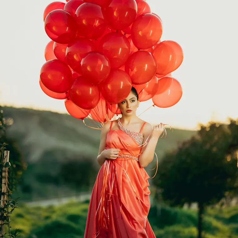Woman in a long coral gown holding a large cluster of red balloons on a hillside.