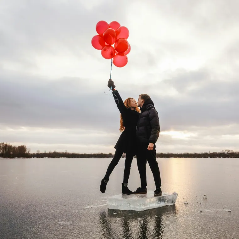 Two people on a lake shore holding a cluster of red balloons at dusk.