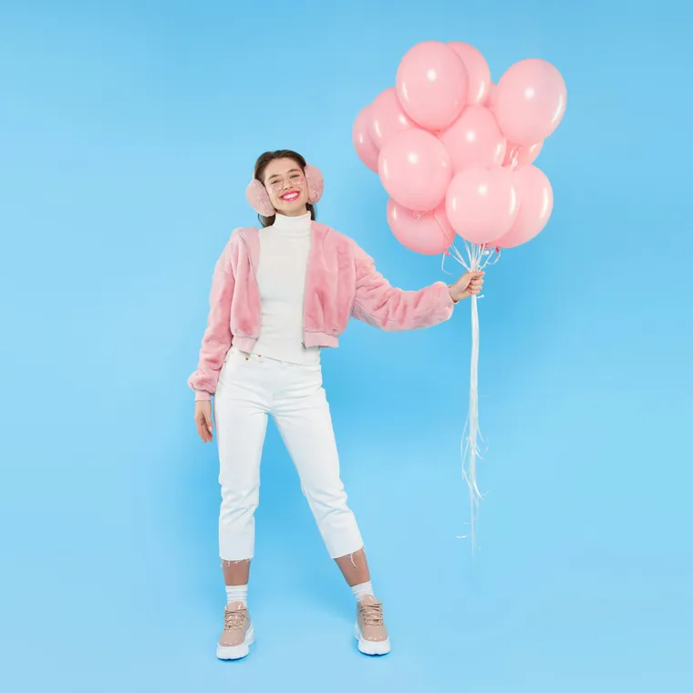 Young girl in a pink jacket holding pink balloons against a blue studio wall.