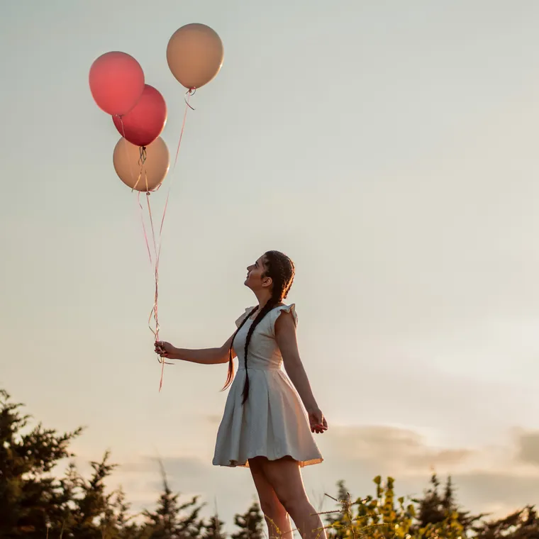 Person in a grey dress holding coral balloons against a sunset sky.
